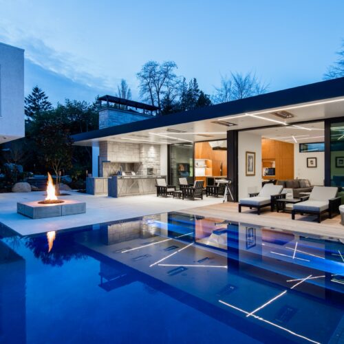 As night falls, the dynamic shapes of the flood edge pool—its curves and crisp angles—take on a new allure, accented by the pool’s deep blue plaster and elegant grey tile band.