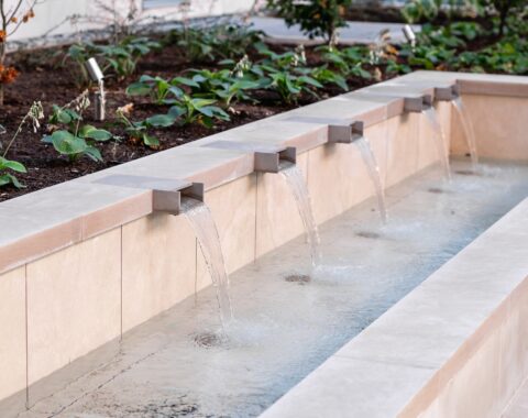 A sleek water feature with evenly spaced weir spouts set amidst modern architectural elements and lush landscaping, providing stunning scenic views. The calming sounds of the waterfall enhance the serene and peaceful atmosphere.
