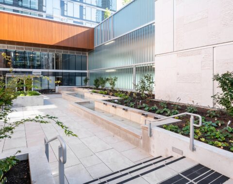 Waterfall feature with a sleek design, surrounded by contemporary architectural elements and landscaped greenery, creating a tranquil atmosphere. People gather to watch and listen to the soothing sounds of the waterfall, enjoying a peaceful and relaxing environment.
